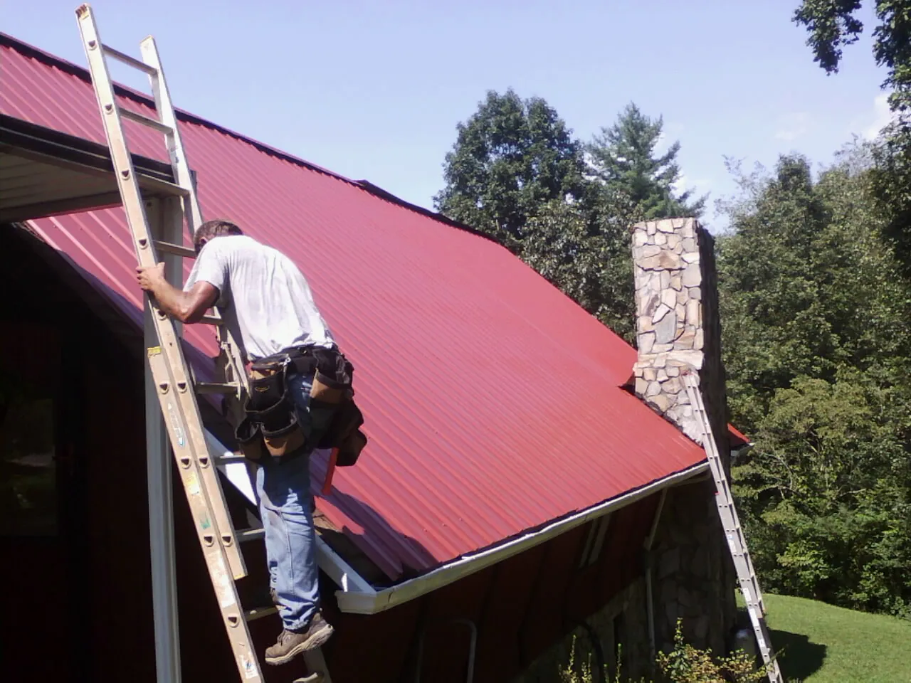 Roofer on a ladder beside a red metal roof and stone chimney — Soffit & Fascia Repair by Roofing Brothers in Delaplane